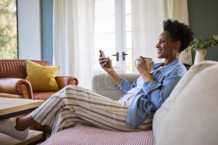 Mature Woman Relaxing On Sofa At Home With Hot Drink Looking At Mobile Phone