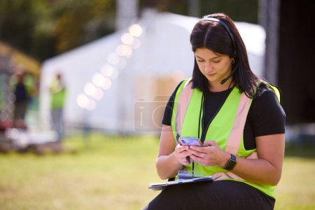 Trabajadora de producción femenina con teléfono móvil que establece el escenario al aire libre para el festival de música o el concierto