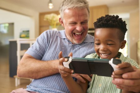Multirassische Familie mit Großvater und Enkel, die gemeinsam auf Handheld-Spielgeräten spielen