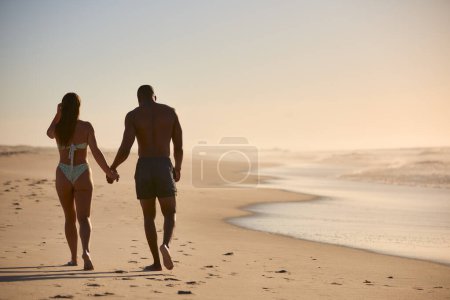 Rear View Of Couple Wearing Swimwear On Vacation Holding Hands Walking Along Beach Shoreline