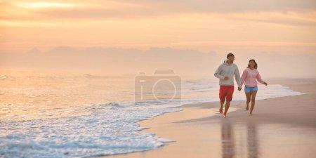 Couple In Casual Clothing On Vacation Holding Hands Running Along Beach Shore At Dawn