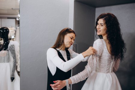 Téléchargez les photos : Femme essayant sur robe de mariée dans un magasin avec des femmes assistant
. - en image libre de droit