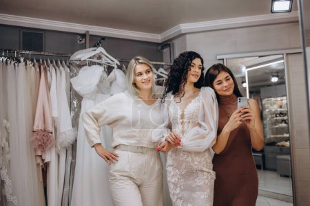 Photo for Women taking photographs of a female friend trying on wedding dress. Women in wedding dress fitting room. - Royalty Free Image