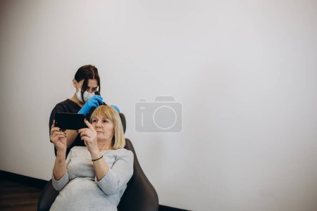 Téléchargez les photos : Un trichologue examine les cheveux d'une patiente âgée avec un trichoscope dans une clinique. Trichoscopie capillaire. Exécute le trichoscope sur le cuir chevelu et regarde le résultat sur l'écran du moniteur. Trichoscopie informatique. - en image libre de droit