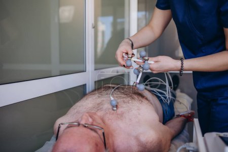 Foto de Cardiólogo examinando paciente mayor con dispositivo Holter en el departamento de cardiología del hospital. La doctora fija sensores para medir el ECG y la presión arterial. Hombre maduro en el sofá de la clínica - Imagen libre de derechos