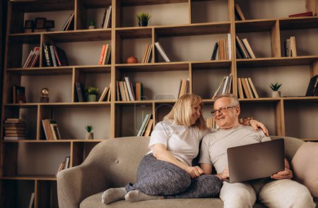 Photo for Mature man and woman using laptop together, looking at screen, older spouses browsing apps, shopping or chatting online, making video call, using bank service, sitting on cozy sofa in living room - Royalty Free Image