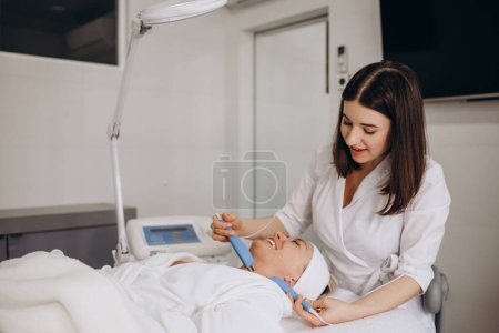 A doctor cosmetologist makes a microcurrent facial therapy to a young woman with a device in a beauty wellness salon.Cosmetology and professional skin care.