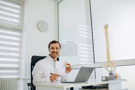 Photo for Young male doctor in uniform and eyeglasses showing vertebra model while speaking about spinal problems during appointment in contemporary office - Royalty Free Image
