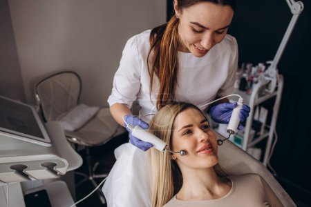 Téléchargez les photos : A doctor cosmetologist makes a microcurrent facial therapy to a young woman with a device in a beauty wellness salon.Cosmetology and professional skin care. - en image libre de droit