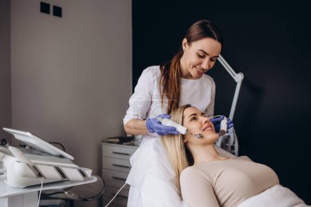 Téléchargez les photos : A doctor cosmetologist makes a microcurrent facial therapy to a young woman with a device in a beauty wellness salon.Cosmetology and professional skin care. - en image libre de droit