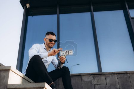 Photo for Funny man throws money. Young happy businessman scatter the dollars in the street near the office center. Money rain, falling dollars. Successful business. - Royalty Free Image