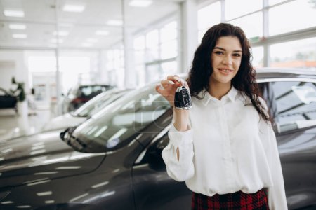 Photo for Happy brunette woman buying new cute car, raising hand up with automatic key and smiling, showroom interior, copy space. Pretty curly lady customer purchasing yellow female auto, getting in. High quality photo - Royalty Free Image