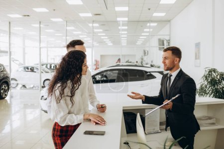 Photo for Sales manager showing car charging station to a young couple, selling electric cars in the showroom. Concept of buying eco-friendly car for family. High quality photo - Royalty Free Image