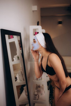 Foto de Una mujer en ropa interior negra con una máscara facial cosmética se mira en el espejo - Imagen libre de derechos