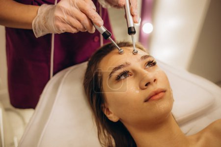Téléchargez les photos : Un médecin cosmétologue fait une thérapie faciale microactuelle à une jeune femme avec un appareil dans un salon de bien-être de beauté. Photo de haute qualité - en image libre de droit