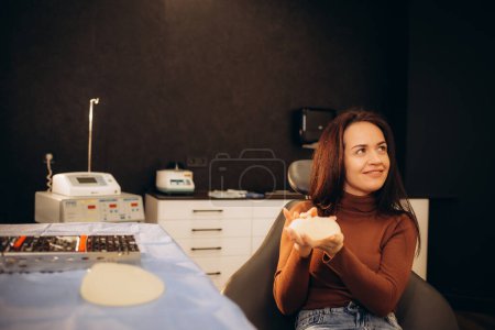 Téléchargez les photos : Gros plan d'une jeune femme souriante heureuse tenant un implant en gel. Travailleur médical joyeux à l'air surpris. Rendez-vous clinique. Médecine moderne et concept de correction corporelle - en image libre de droit
