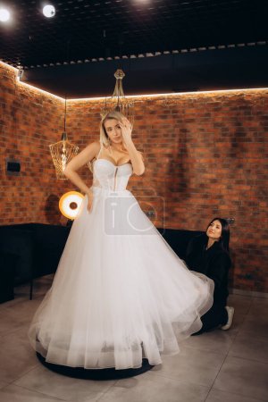 Téléchargez les photos : Jeune belle mariée caucasienne va acheter robe de mariée par designer dans le salon, essayez-le et regardez miroir. Photo de haute qualité - en image libre de droit