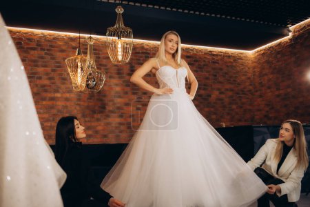 Téléchargez les photos : Couturière aidant la mariée à mettre sa robe de mariée dans le magasin de vêtements. Photo de haute qualité - en image libre de droit