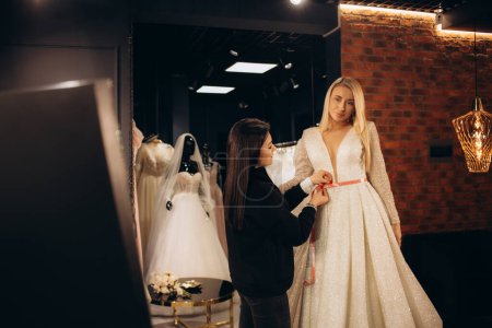 Téléchargez les photos : Belle mariée s'habiller par sa meilleure amie dans son jour de mariage et choisir une robe de mariée dans la boutique et l'assistante de magasin l'aide. Photo de haute qualité - en image libre de droit