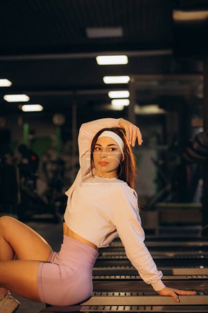 Young sexy fitness girl in sport wear relaxing on the treadmill in the gym, woman exercise working out sitting on runnig machine. High quality photo