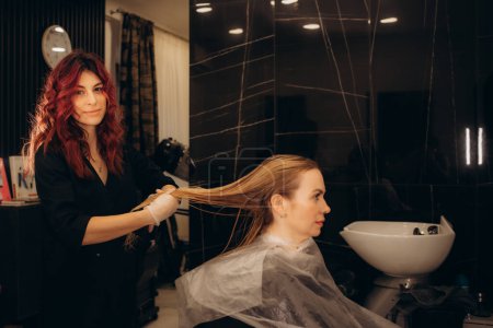 Téléchargez les photos : Une coiffeuse teint les cheveux d'une jeune femme caucasienne avec une brosse et une feuille dans un salon de beauté. Photo de haute qualité - en image libre de droit