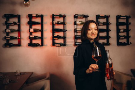 Focused female wine expert smells wine into glass, holding bottle and reads label with characteristics of wine, the year of manufacture, region and the degree of acidity of the alcoholic beverage.