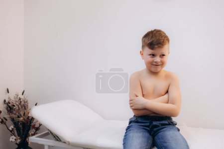 A little boy without a T-shirt sits on a couch in a hospital