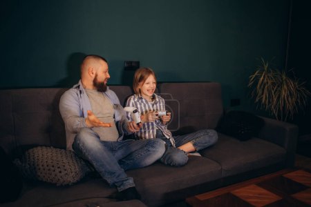 Happy father and little daughter playing video game at home, smiling preschool girl and young dad holding gamepads controllers, sitting on couch in living room, having fun with gadgets