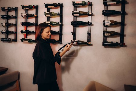 Cheerful caucasian female sommelier opens a bottle of wine with a corkscrew, dressed in classic strict clothes, prepares for wine tasting while in wine cellar among many different wines.