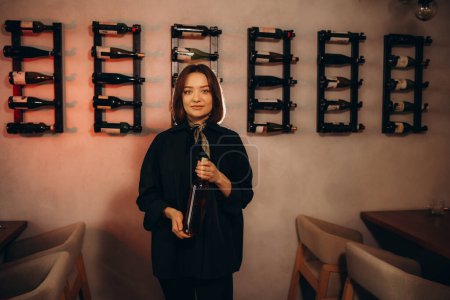 Cheerful caucasian female sommelier opens a bottle of wine with a corkscrew, dressed in classic strict clothes, prepares for wine tasting while in wine cellar among many different wines.
