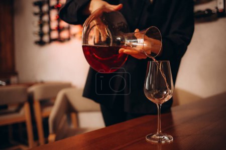 Female sommelier pouring red wine to decanter for decanting an alcoholic beverage, standing in cellar against shelf with a many and various drinks. 