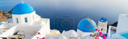 Biała dzwonnica kościelna, niebieskie kopuły i kaldera wulkaniczna z morskim krajobrazem, piękne szczegóły wyspy Santorini z kwiatami, Grecja