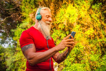 Photo for Senior man with white beard doing running outdoor on the nature, holding phone and choosing music - Royalty Free Image