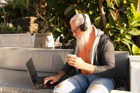 Photo for Senior smiling man with laptop and earphones, relaxing on cafe or home terrace - Royalty Free Image