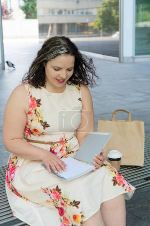 Foto de Retrato de mujer de 40 más tamaño con sentado en un banco al aire libre en una ciudad y haciendo avisos en el cuaderno - Imagen libre de derechos