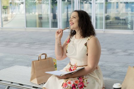 Foto de Retrato de mujer de 40 más tamaño con sentado en un banco al aire libre en una ciudad y haciendo avisos en el cuaderno - Imagen libre de derechos