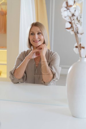 Skin care. Beautiful senior woman looking at herself at mirror doing face fitness - self face massage, sitting in front of mirror at home, making beauty routine procedures