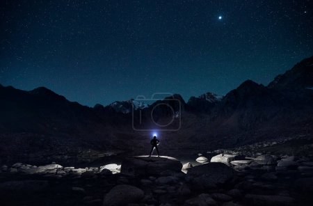 Photo for Tourist man in silhouette with light beam of headlamp near lake in the mountains under night sky with stars. - Royalty Free Image