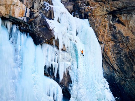 Aerial drone view of athlete in orange jacket Ice climbing at big frozen waterfall in Barskoon gorge the mountain valley in Kyrgyzstan