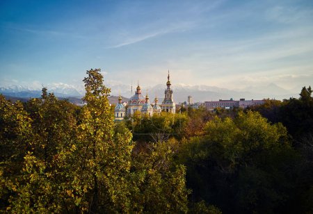 Aerial drone view of the Ascension Cathedral Russian Orthodox church and snow mountains at background in Panfilov Park with tree at foreground in Almaty city, Kazakhstan