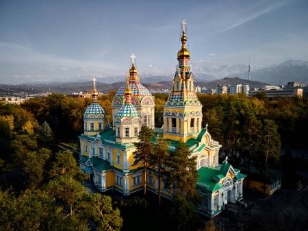 Aerial drone panorama of the Ascension Cathedral Russian Orthodox church and snow mountains at background in Panfilov Park against sunset sky in Almaty city, Kazakhstan