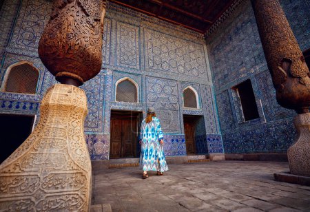 Woman tourist in ethnic dress at Inner Yard of blue mosaic palace hall in Tash Hauli with wooden column at ancient city Khiva in Uzbekistan.