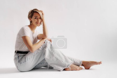 Téléchargez les photos : Corps complet de jeune femme pieds nus assise sur le sol tout en se relaxant sur fond blanc - en image libre de droit
