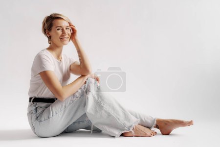Téléchargez les photos : Corps complet de jeune femme pieds nus assise sur le sol tout en se relaxant sur fond blanc - en image libre de droit