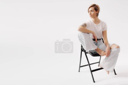 Téléchargez les photos : Corps entier de jeune femme pieds nus assise avec jambe relevée sur chaise tout en touchant le genou avec la main et isolé sur fond blanc - en image libre de droit