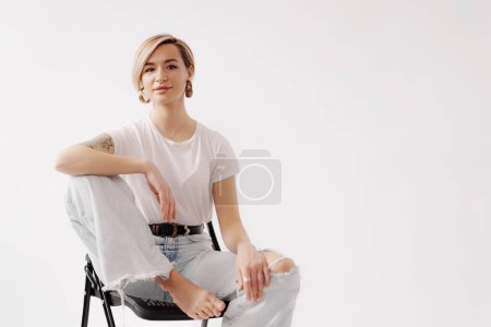 Full body of barefoot young female sitting with raised leg on chair and looking at camera while touching knee with hand and isolated against white background