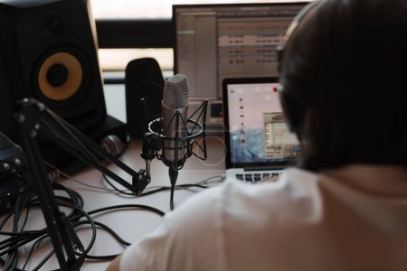 Side view of focused male musician in white t shirt and headphones typing on laptop while recording podcast in modern studio