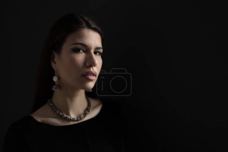 Photo for Portrait of attractive young female model wearing black dress with necklace and earrings looking at camera against dark background in darkness - Royalty Free Image