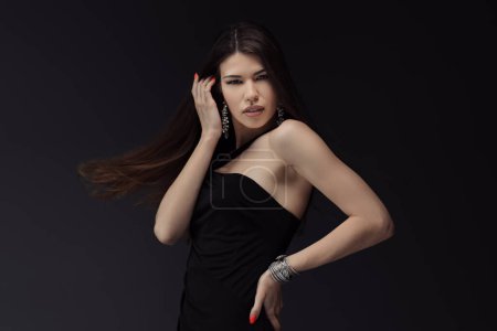 Photo for Young attractive female in black dress touching hair and posing for photo session against dark background looking at camera with toothy open mouth - Royalty Free Image
