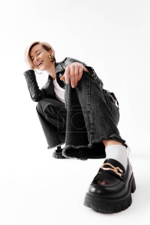Full body of young female in leather jacket and boots sitting on haunches on floor while hand touching hair against white background